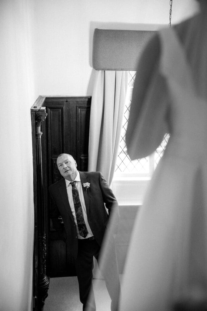 Father of the bride looks up the stairs at his daughter in her wedding dress at the River Barn