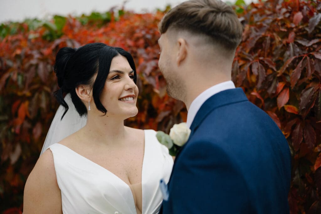 Bride and groom smile at each other with deep red foliage in the background, outside the River Barn, Poulton--le-Fylde, by Nessworthy Pictures