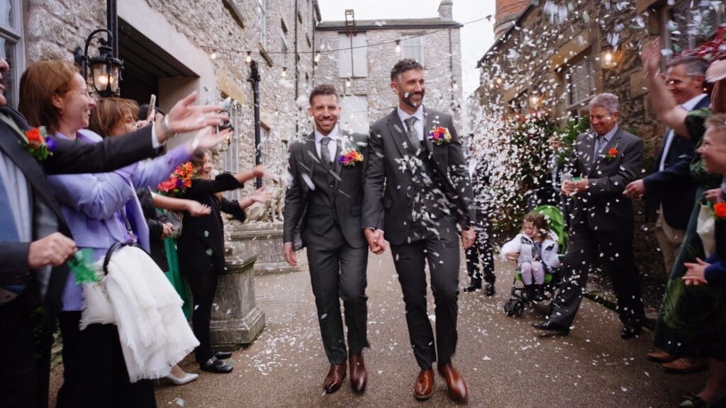 Still showing the couple walking towards the camera holding hands as their guests shower them with confetti. Taken from Andi & Davide's wedding film, captured by Holmes Mill wedding videographer, Nessworthy Pictures.